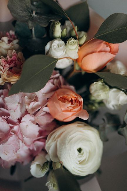 A close-up of a floral arrangement featuring peach-colored roses, white lisianthus, and pale pink hydrangeas, accented with large dark green eucalyptus leaves. The flowers are arranged in a bouquet-st