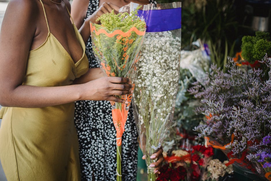 A person wearing a yellow sleeveless dress is holding a large, conical bouquet wrapped in orange and clear cellophane, filled with green and white filler flowers. They are standing at a flower shop or