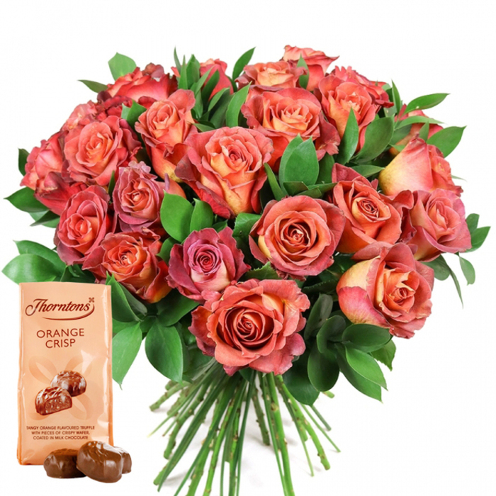 Dominating the centre of this elegant image is a generous, rounded bouquet of fresh roses, their colours flowing from delicate, golden peach in tight centres to vivid coral, salmon, and rich pink at the petal edges. This remarkable bicolour effect creates visual depth and movement reminiscent of Lower Clapton's evolving landscape. The roses, tightly clustered for a lush, full effect, are surrounded by abundant greenery-likely ruscus or salal-whose verdant tones ground the arrangement and highlight the warmth of the blooms. Each petal appears velvety and soft, catching natural light to enhance the flowers' brilliance, as though they've been freshly gathered from nearby gardens. Anchoring the lower-left foreground is a Thorntons Orange Crisp chocolate pouch, its light cream packaging elegantly displaying the brand mark and bold "Orange Crisp" label. Below, charming illustrated truffles and descriptive text invite closer inspection, depicting the chocolates as "tangy orange flavoured truffles with pieces of crispy wafer, coated in milk chocolate." Completing the tableau are two realistic, glossy milk chocolate truffles, carefully placed in front of the packet, their surface reflecting the ambient light and suggesting irresistible indulgence. Long, straight rose stems peek out beneath the floral canopy, confirming the bouquet's freshness and care. The white background ensures every vivid detail stands out, making this composition the perfect expression of gratitude, love, or celebration-ideal for occasions in Lower Clapton, from milestone birthdays to anniversaries, or simply as a heartfelt gesture of appreciation.